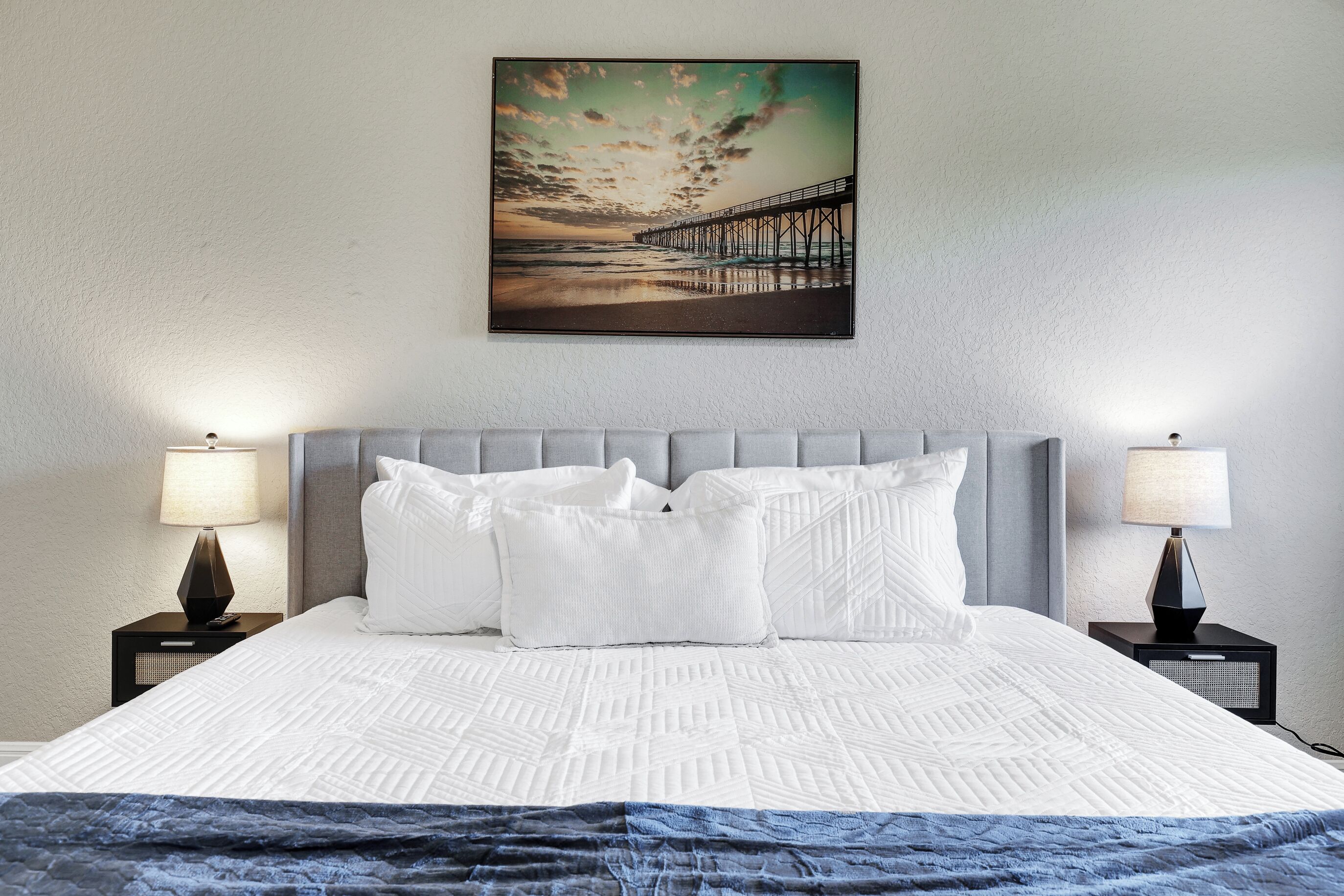 A neatly made bed with white linens, two bedside tables with lamps, and a painting of a pier on the wall.