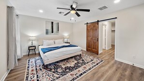 A bedroom with a bed, a ceiling fan, a nightstand, a lamp, a wooden door, and a patterned rug.