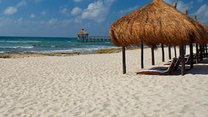 Une plage avec des parasols en chaume et des chaises longues, une jetée en arrière-plan et un ciel bleu dégagé.