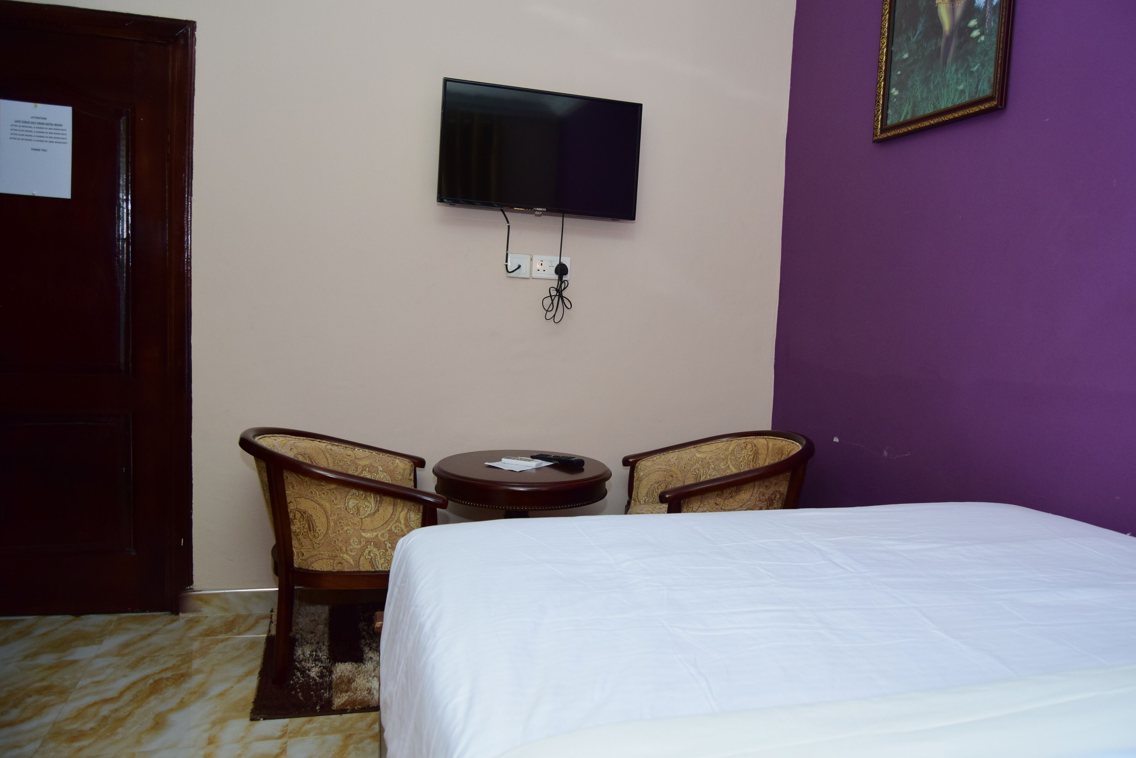 A hotel room with a bed, a television mounted on the wall, and two armchairs around a small table.