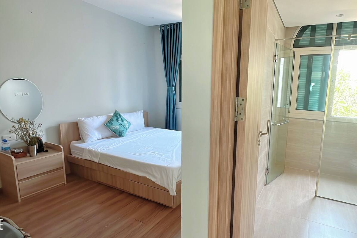 A bedroom with a bed, bedside table, and a mirror. There is a bathroom with a glass shower enclosure and a toilet.