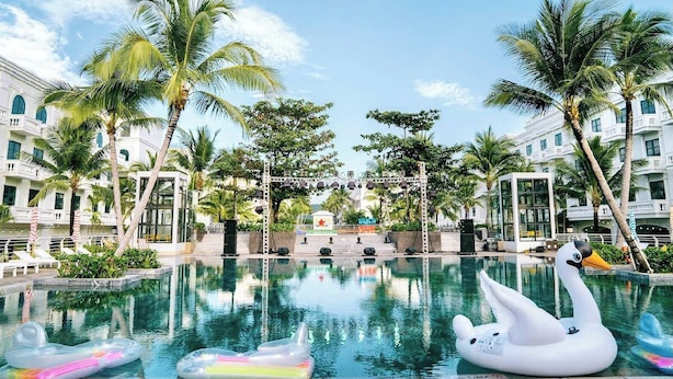 A swimming pool with a swan float, palm trees, and a building with balconies. - The Costis Homestay & Hotel  (Phu Quoc)