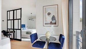 A modern hotel room with a bed, a dining area with two blue chairs and a table, a large abstract painting on the wall, and a view through a glass door. - Studio Sleek & Refined - Levallois-Perret (Levallois-Perret)