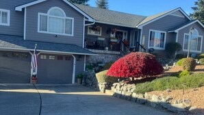 A two-story house with a garage, a front porch with outdoor seating, and a well-maintained garden with red foliage.