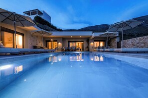A modern pool area with lounge chairs and umbrellas, surrounded by a stone wall and a mountain backdrop.