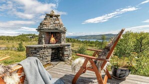 A stone fireplace with a fire, a wooden deck, a chair, and a potted plant.