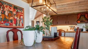 A dining area with a wooden table, chairs, and potted plants. There is a kitchen in the background with wooden cabinets and a hanging knife rack.