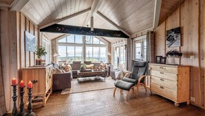 A cozy, well-lit room with wooden walls and a high ceiling. It features a large window offering a view of the outdoors, a comfortable seating area with a sofa and chairs, and a wooden cabinet with drawers.