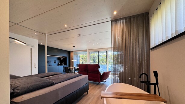 Une chambre moderne avec un grand lit, une table de chevet en bois, une chaise rouge, une baignoire et un coin cuisine visible à travers une porte coulissante.