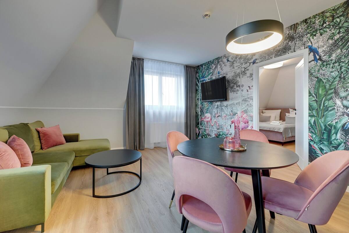 A modern living room with a green sofa, pink chairs, a round table, and a TV mounted on the wall.