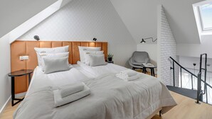 A modern bedroom with a sloped ceiling, a wooden headboard, a bed with white linens, a small side table, a chair, and a balcony with a railing.
