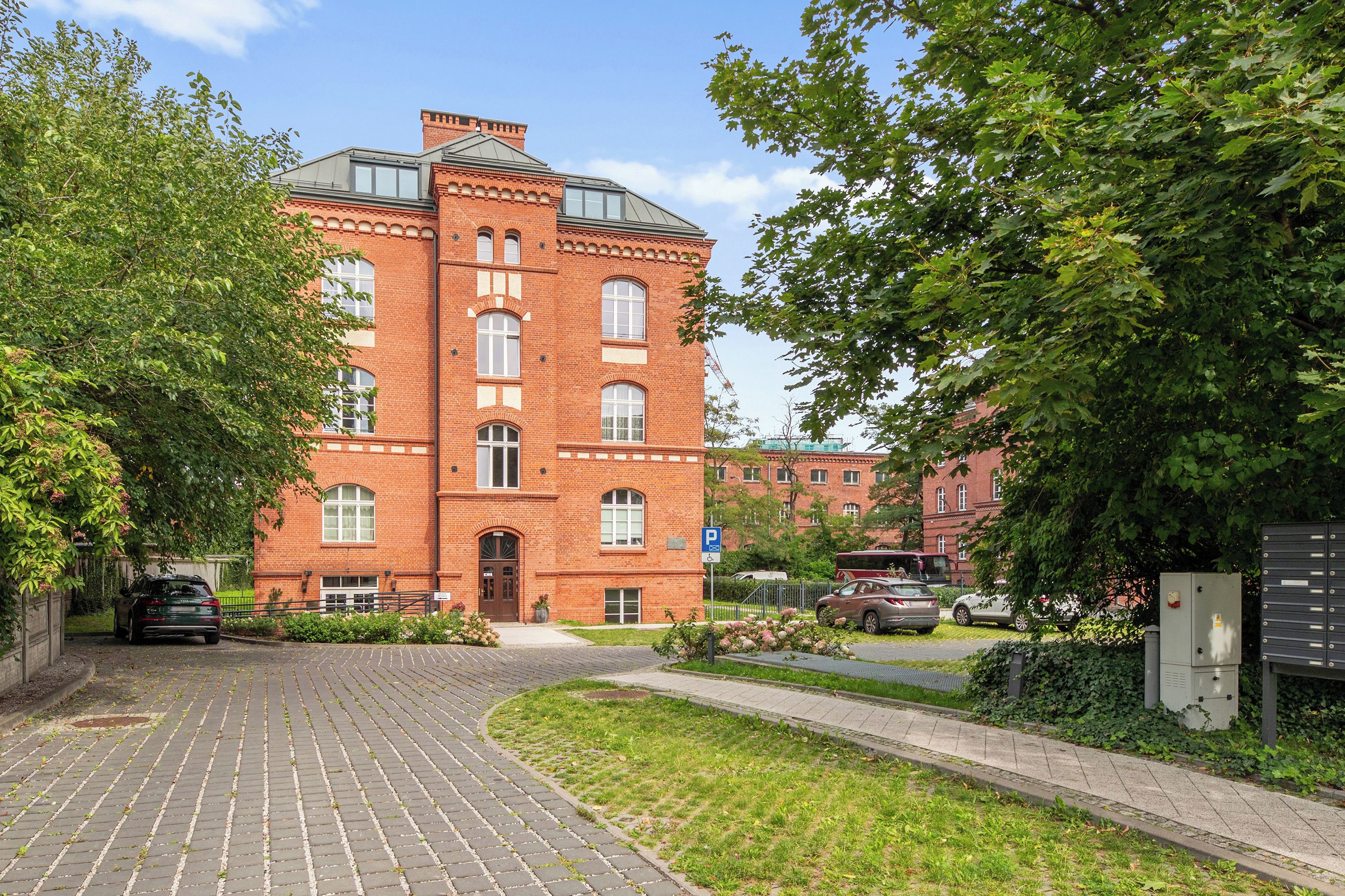 A red brick building with a central entrance, a paved driveway, and a parking area with cars.
