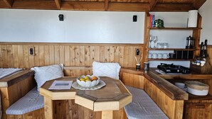 A wooden cabin interior with a dining table, two cushioned benches, a fruit bowl, and a shelf with kitchenware.