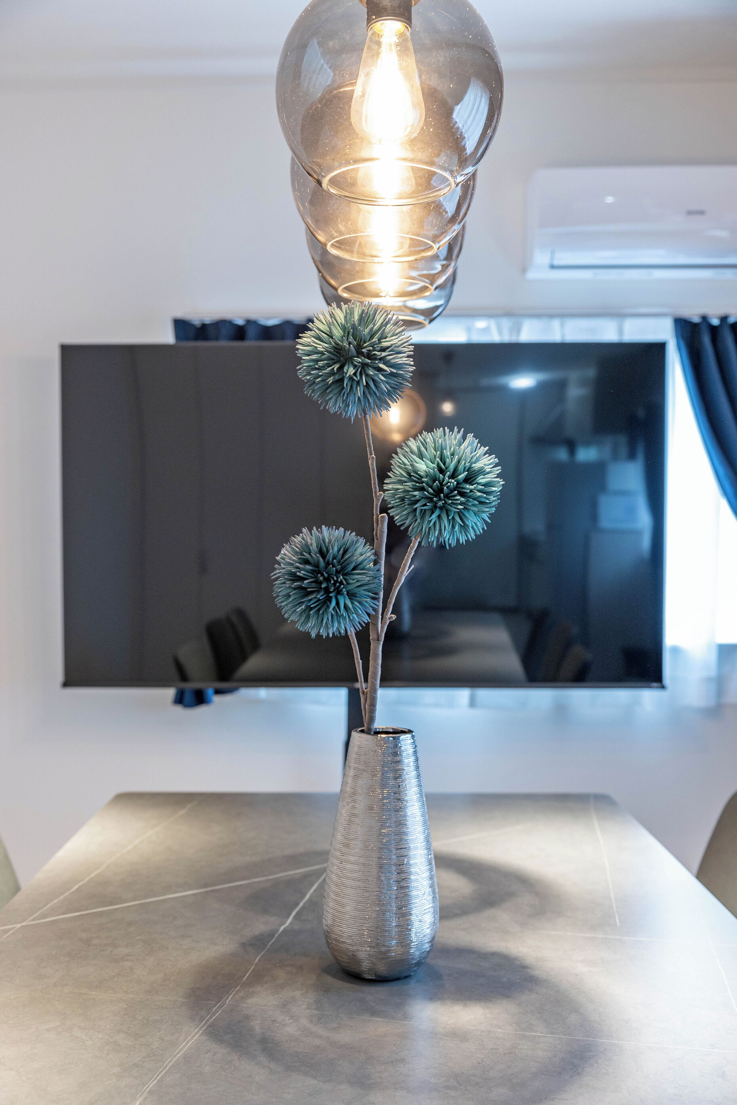 A modern interior with a table lamp, a vase with decorative spheres, and a large mirror reflecting a room with a dark-colored sofa and a television.