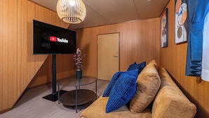 Ein modernes Wohnzimmer mit einem Flachbildfernseher, der das YouTube-Logo anzeigt, einer Holzwand, einem Glastisch und einem braunen Sofa mit blauen Kissen sowie einer dekorativen Vase.