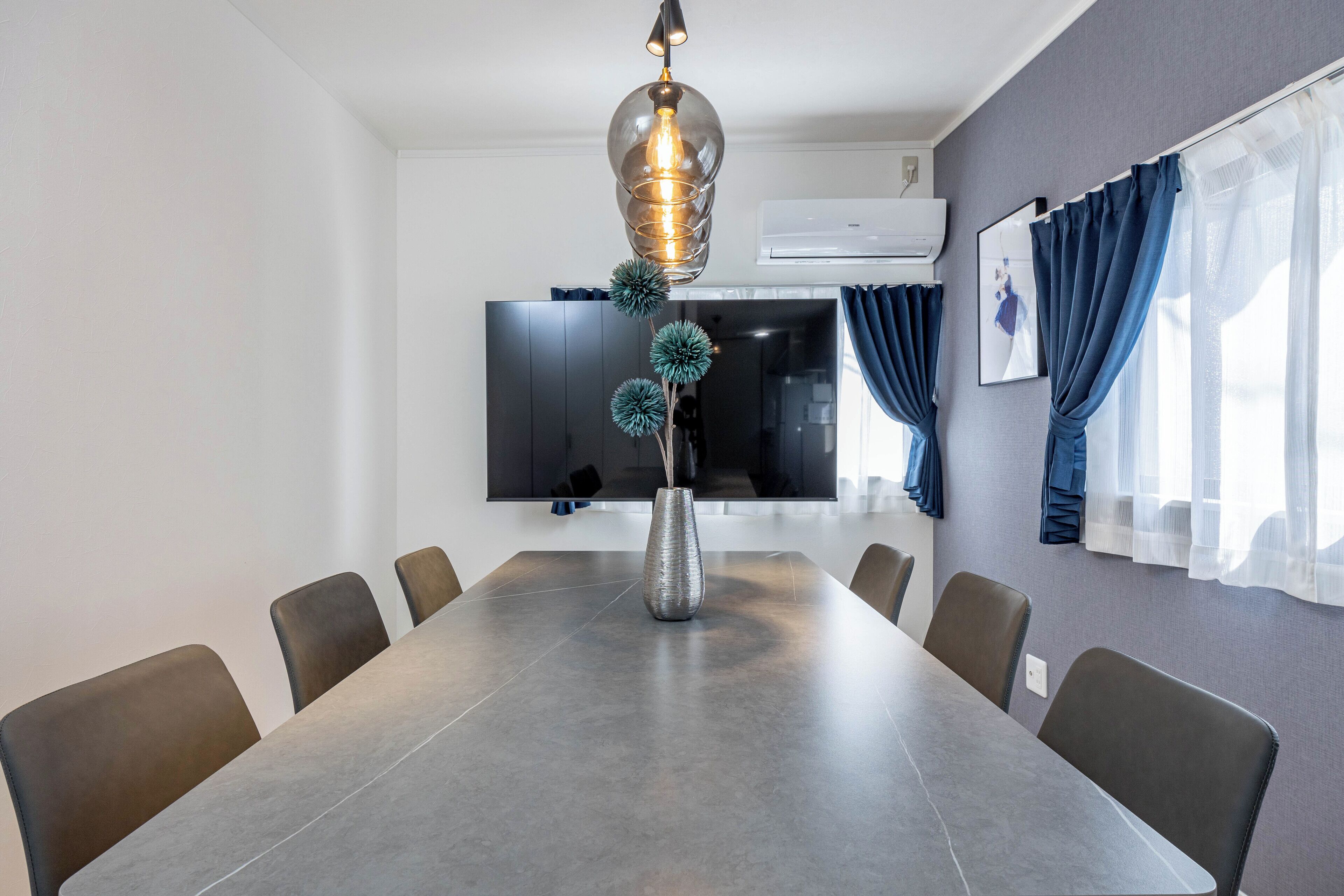 A conference room with a long table, chairs, a decorative vase, a modern pendant light, a flat-screen TV, and a window with curtains.