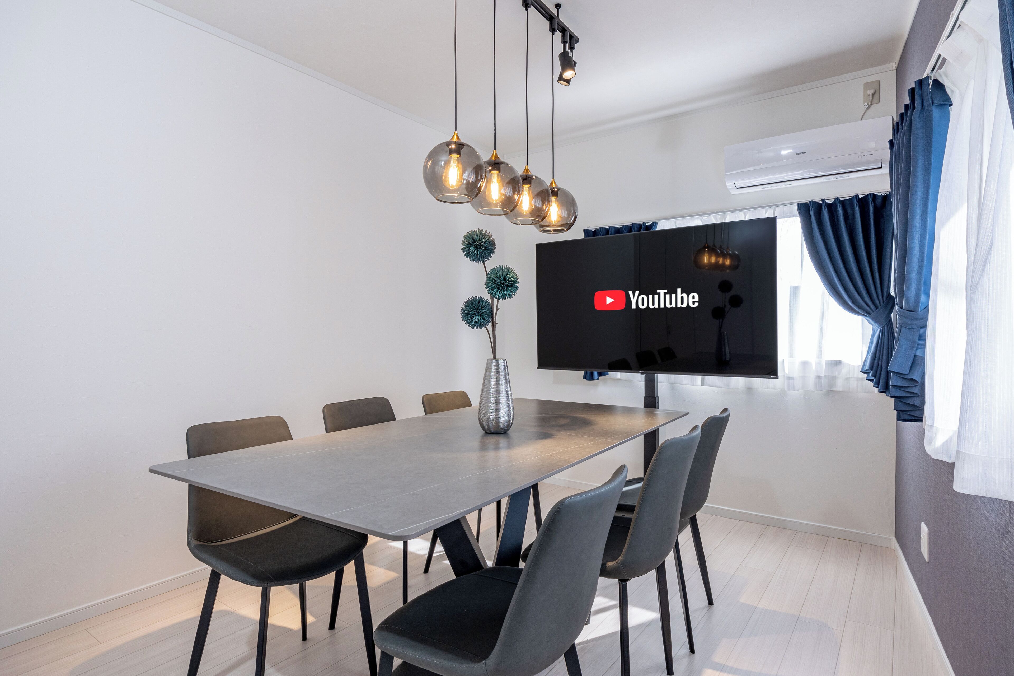 A modern dining area with a long table, six chairs, a vase, and a flat-screen TV mounted on the wall.