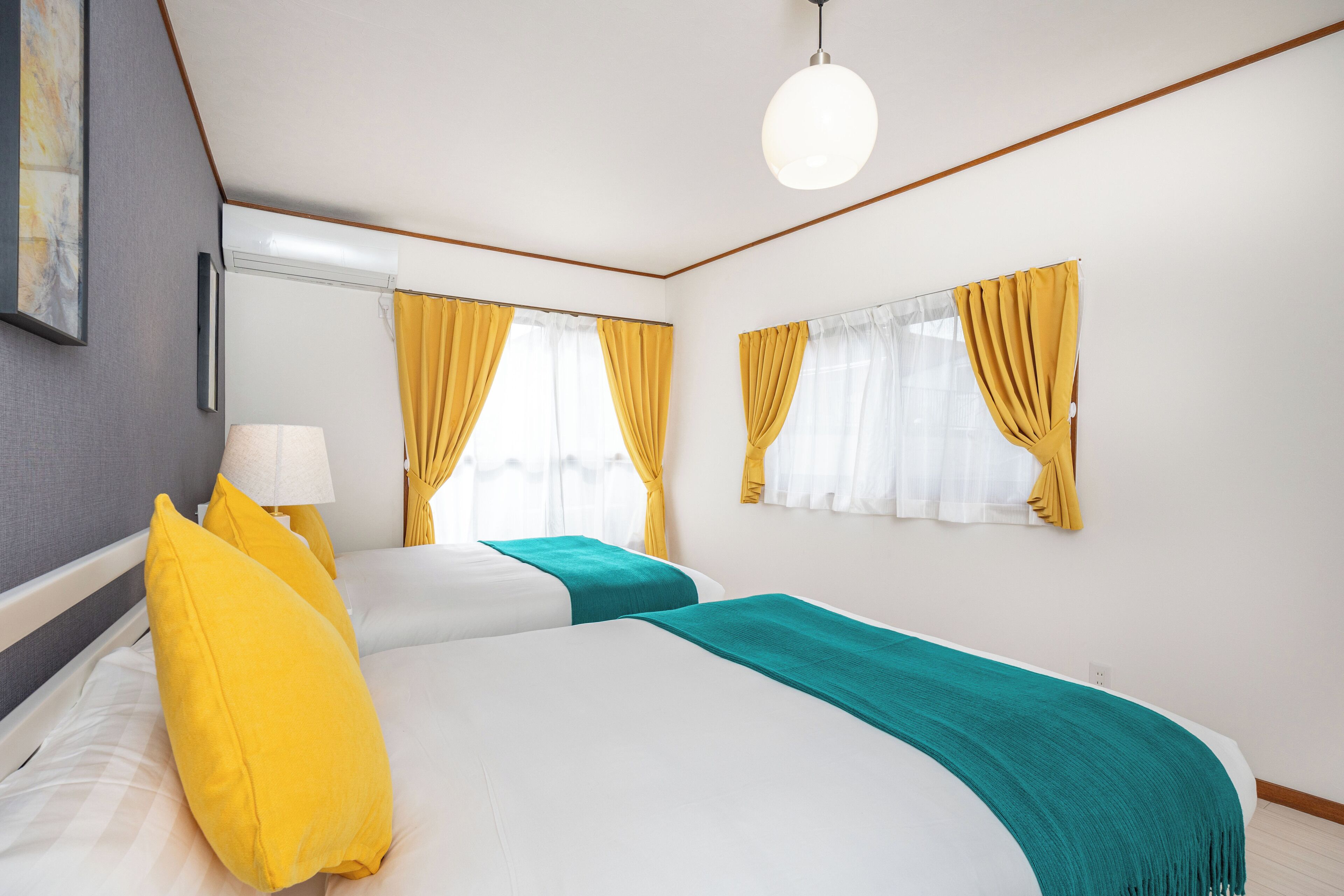 Ein Hotelzimmer mit zwei Betten, gelben Vorhängen, einer türkisfarbenen Tagesdecke und einer weißen Deckenleuchte.