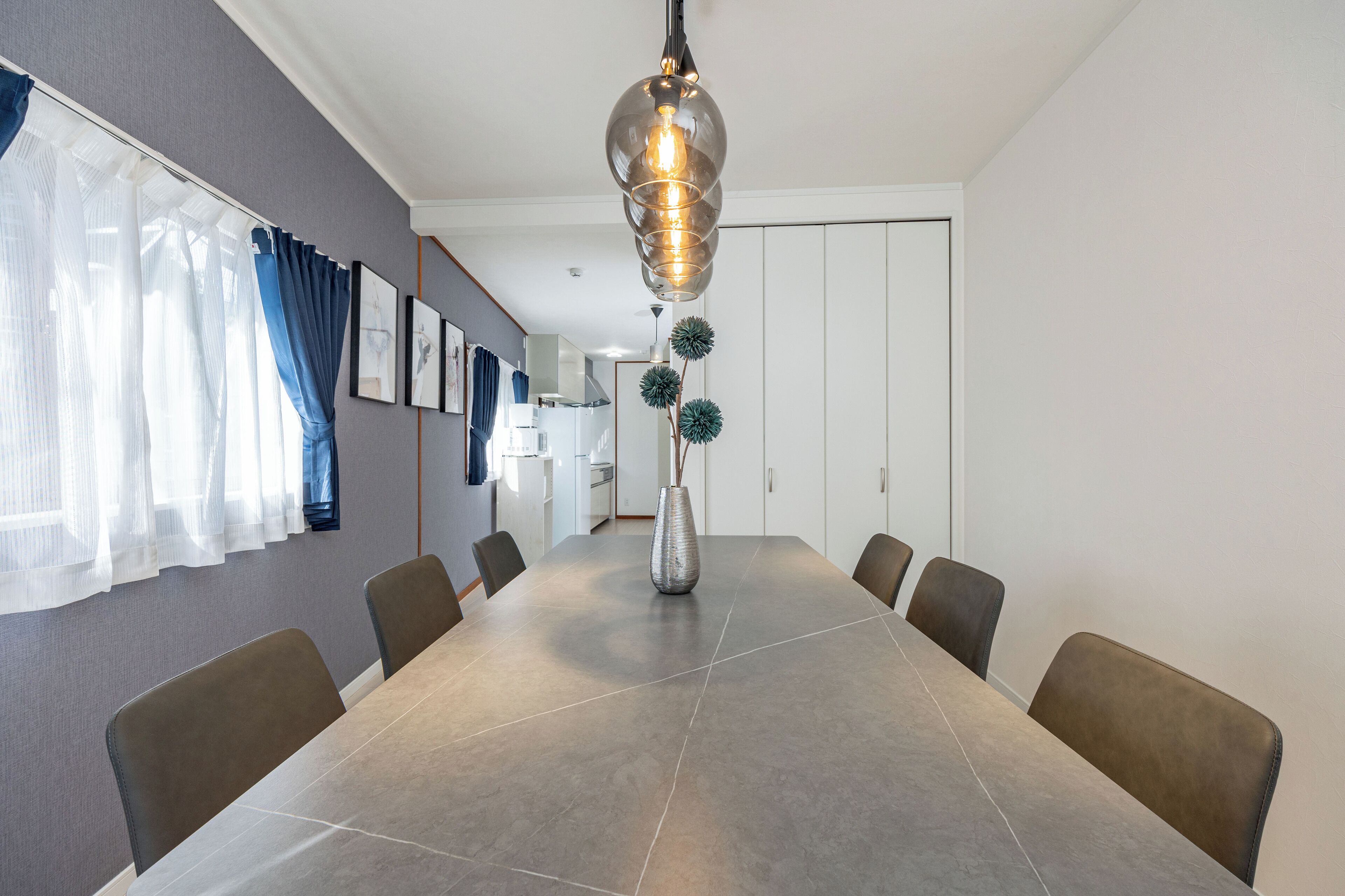 A modern dining area with a large table, chairs, a hanging light fixture, and a vase with decorative plants.