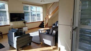 A modern living room with a flat-screen TV, grey sofas, wooden flooring, a staircase, and a built-in fridge. - Tranquil Retreat in Gorebridge (Gorebridge)