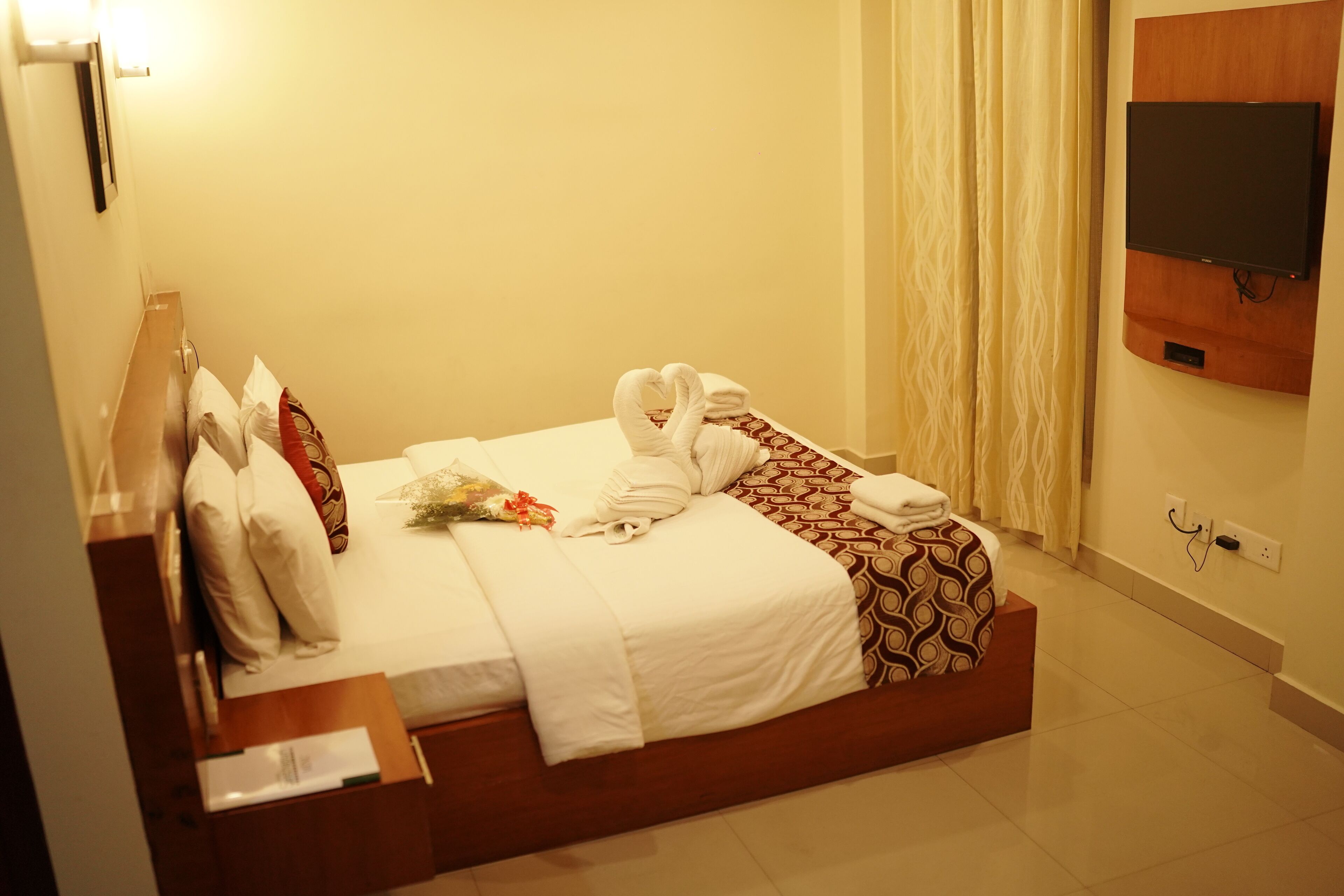 A hotel room with a neatly made bed, a flat-screen TV mounted on the wall, and a bedside table with a lamp.
