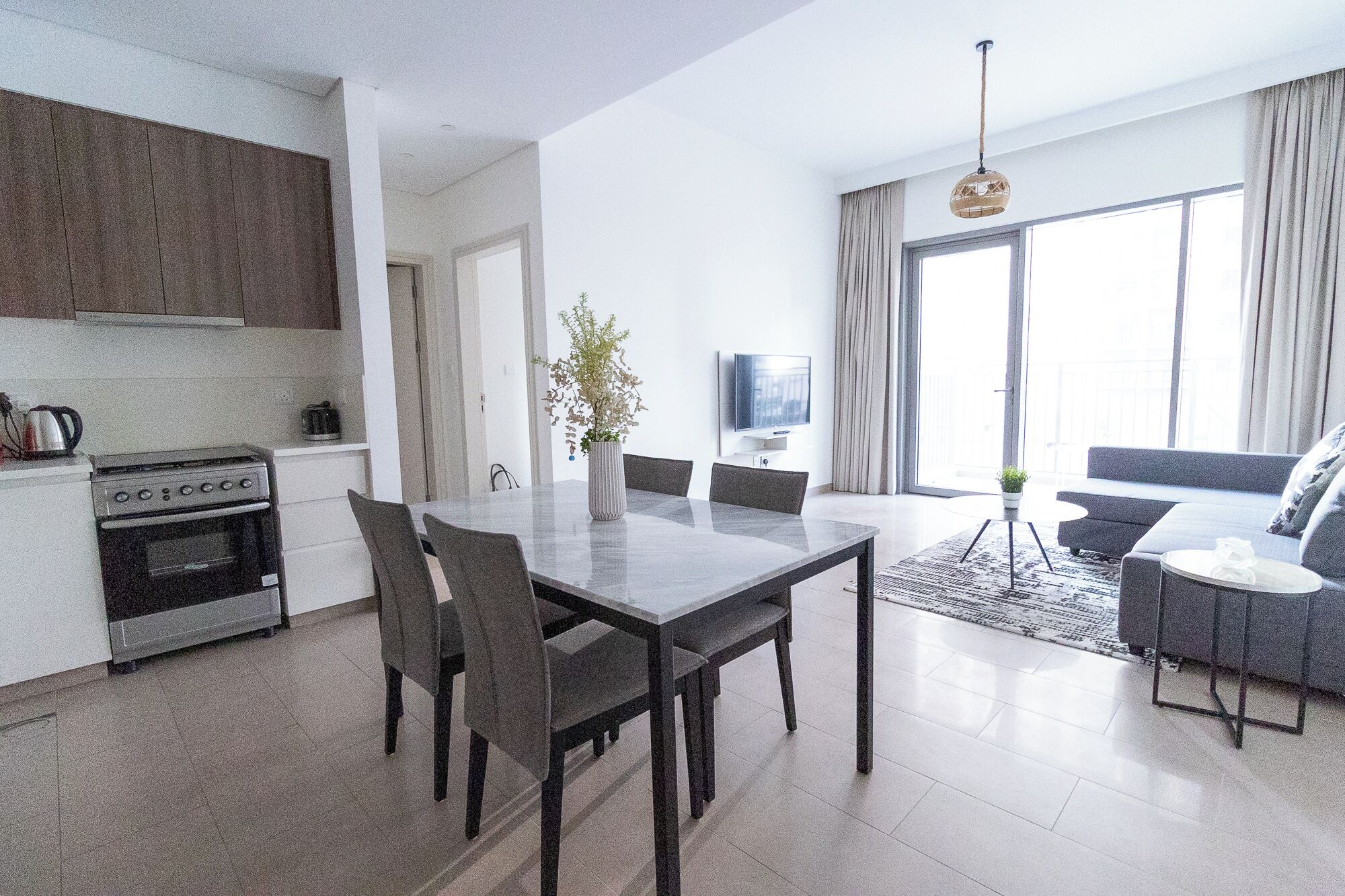 Een moderne keuken met een fornuis en kastjes, een eethoek met een tafel en stoelen, een zithoek met een bank en een televisie, en grote ramen met gordijnen.