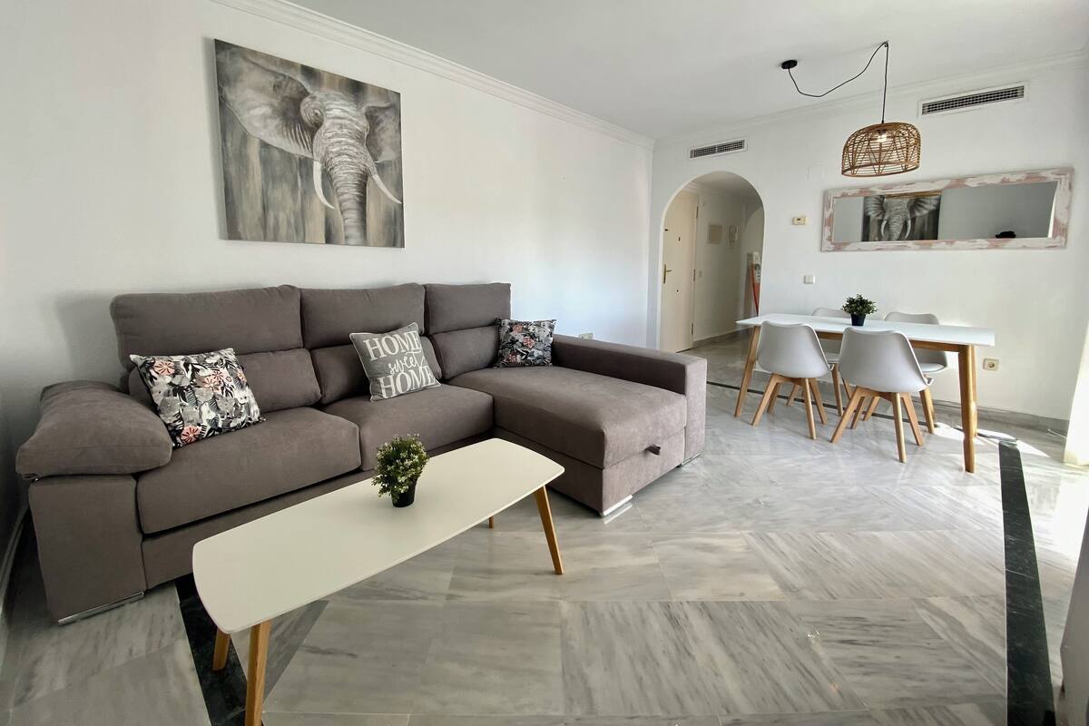 Un salon moderne avec un canapé d’angle gris, une table basse blanche et un coin repas comprenant une table blanche et des chaises grises.