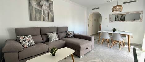 A modern living room with a grey sectional sofa, a white coffee table, and a dining area with a white table and grey chairs.