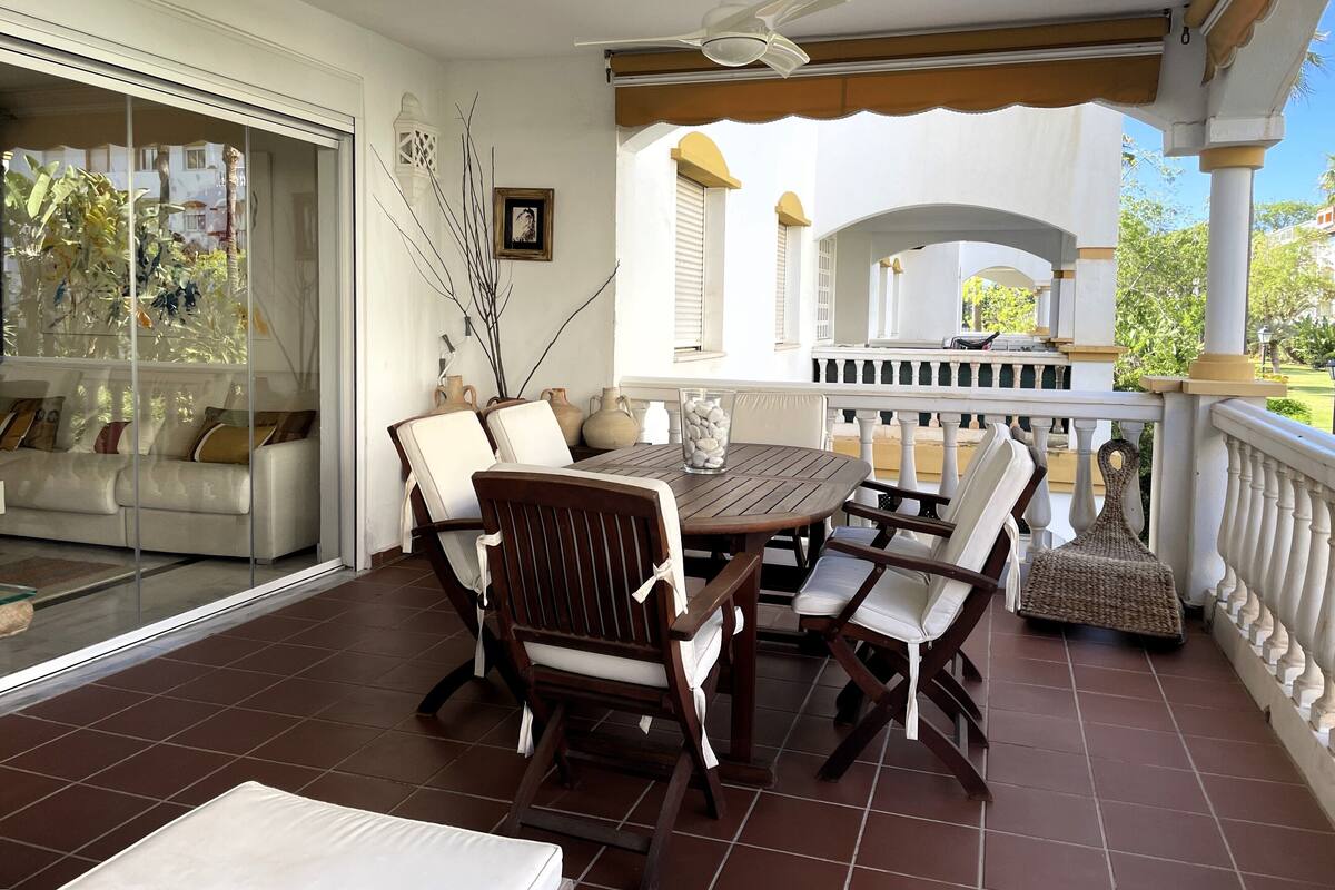 Une terrasse couverte avec une table et des chaises en bois, un canapé blanc et un vase décoratif.