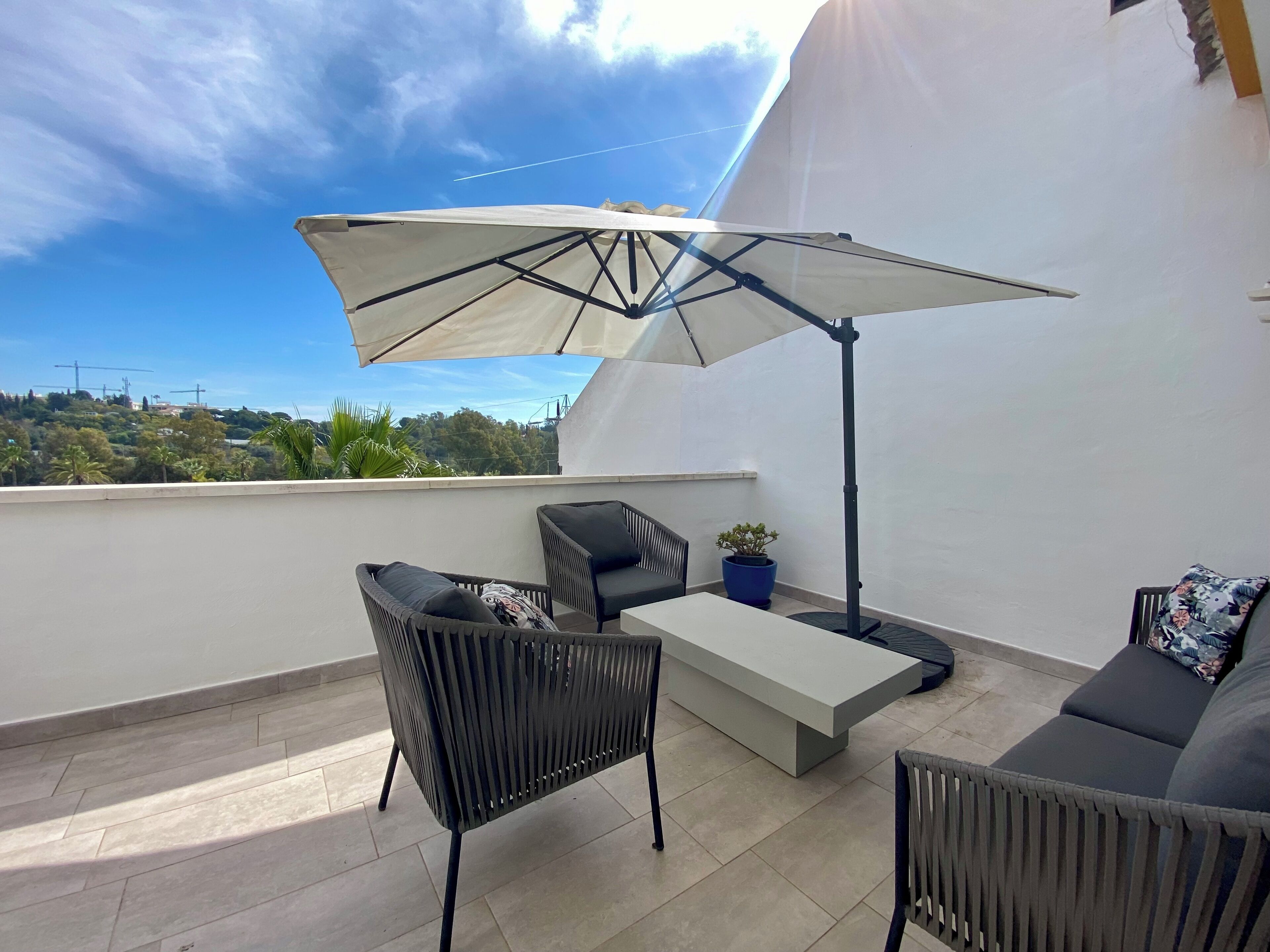 Une terrasse sur le toit avec un parasol blanc, deux chaises en osier noir, un canapé gris et une petite plante en pot.