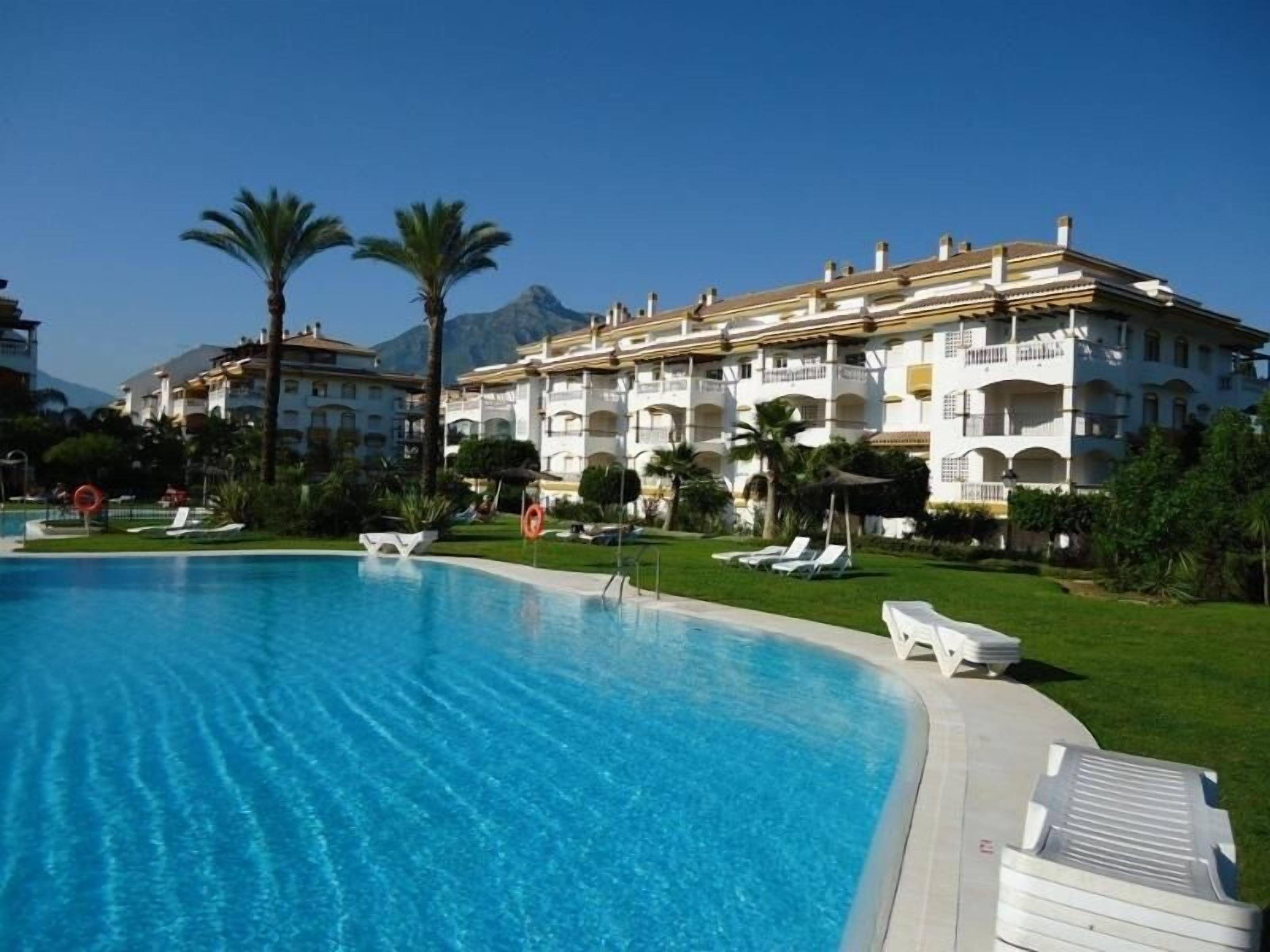 A swimming pool with lounge chairs, palm trees, and a multi-story building with balconies.
