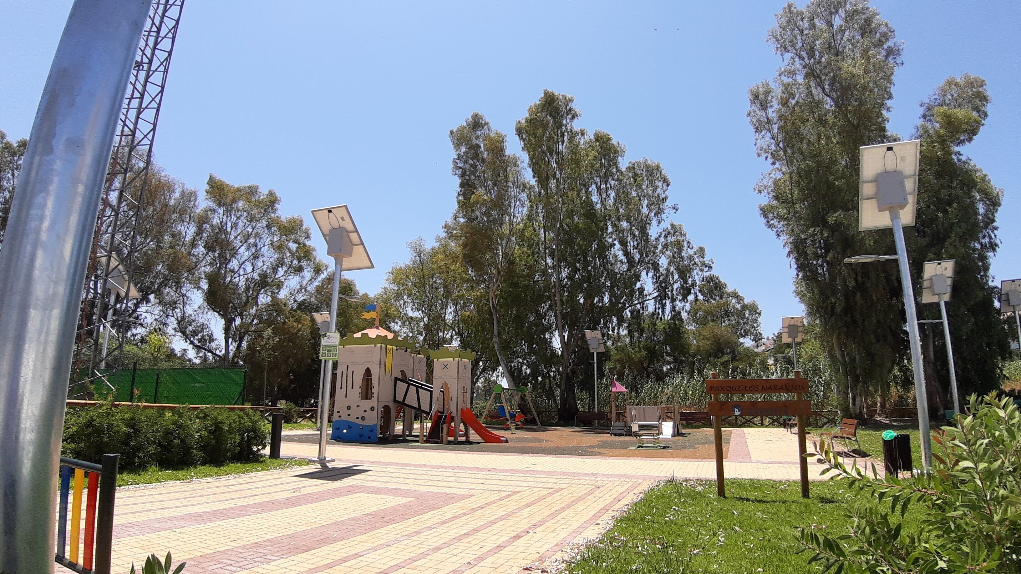 Un parque infantil con tobogán y estructuras para escalar, rodeado de árboles y una zona pavimentada.