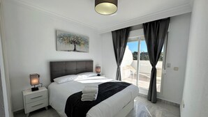 Una camera da letto moderna con un letto grande, un comodino con una lampada, una luce a soffitto, un quadro al muro e vista all'esterno attraverso una porta scorrevole.