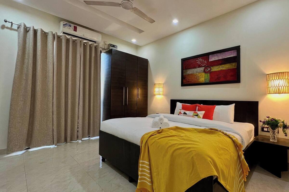 A hotel room with a bed, a wardrobe, a ceiling fan, an air conditioning unit, a painting, and two wall-mounted lamps.
