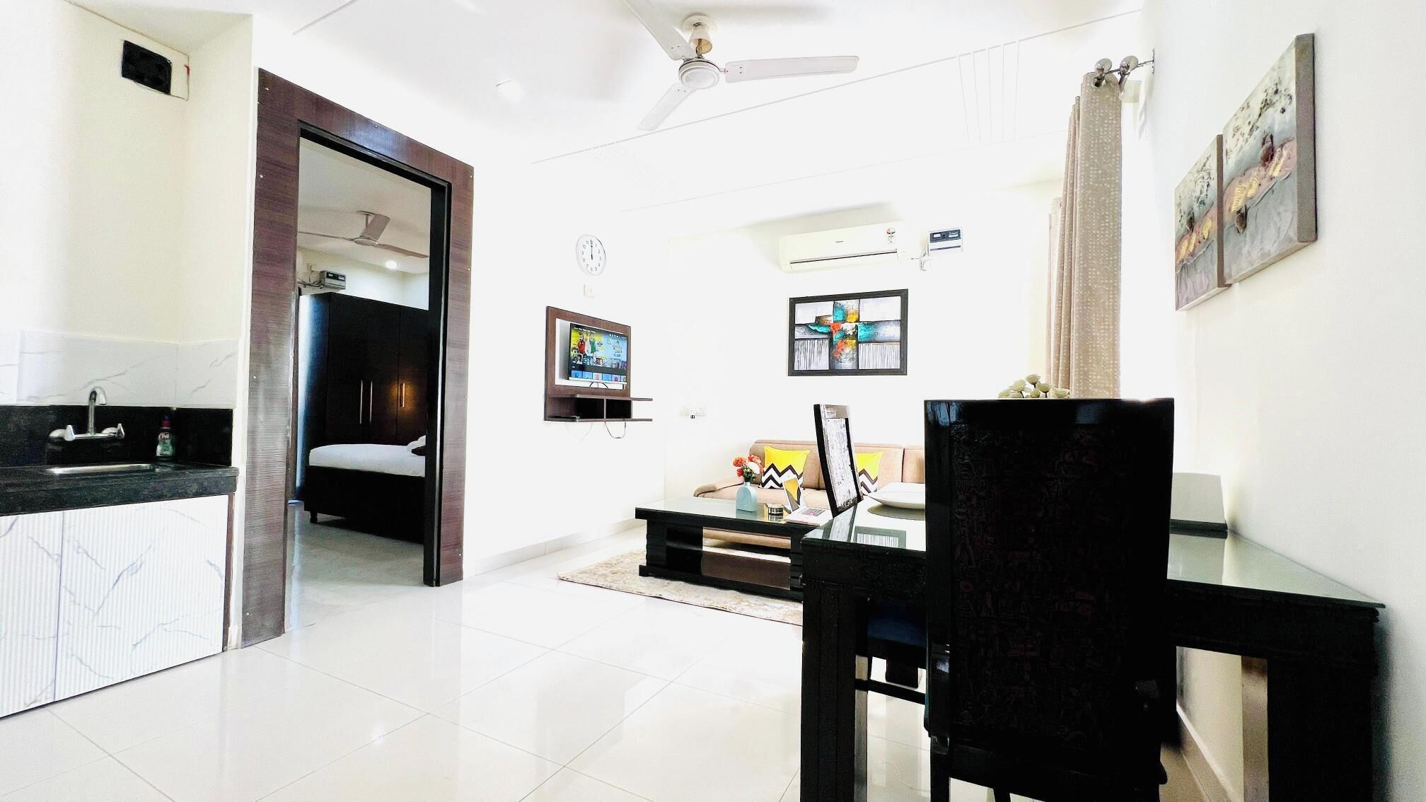A modern living room with a sofa, a coffee table, and a TV. There is a dining table with chairs and a kitchen area with a sink and cabinets.