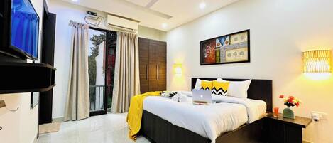 A hotel room with a bed, bedside table, TV, air conditioning unit, and a painting on the wall.
