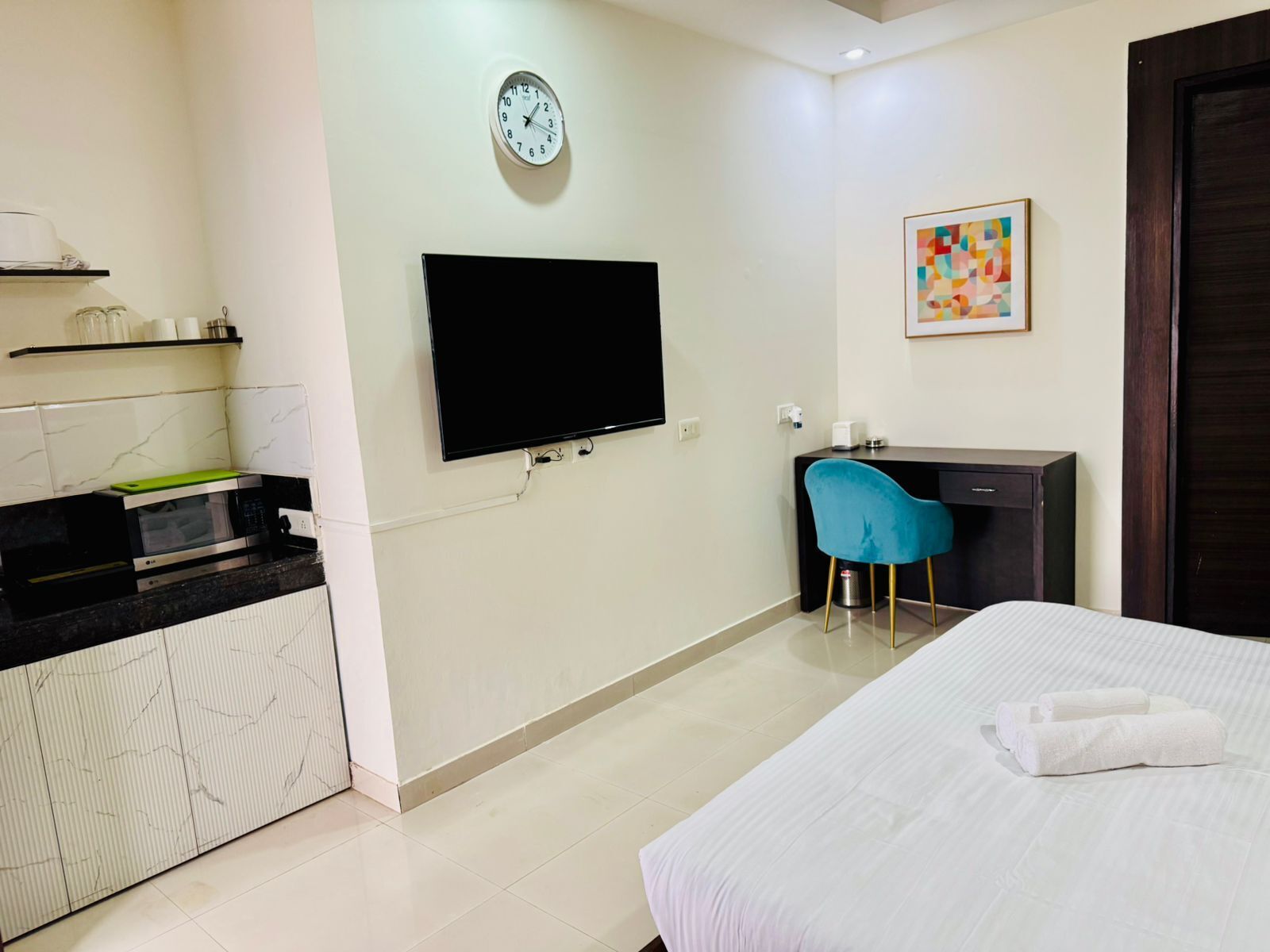 Habitación de hotel con cama, escritorio, televisión, reloj y kitchenette.