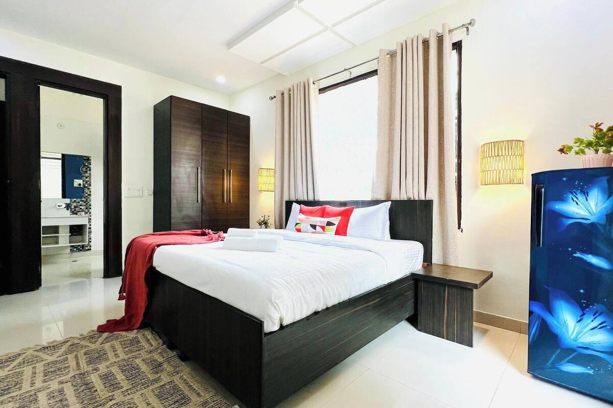 A modern hotel room with a large bed, dark wooden wardrobe, a small bedside table, a decorative wall lamp, and a large mirror.