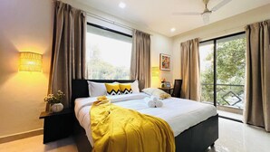 A hotel room with a bed, a nightstand, a lamp, a window with curtains, and a balcony view. - Bluo Stays - DLF Cybercity (Gurugram)