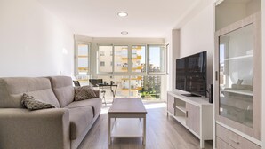 Una sala de estar moderna con un sofá, una mesita de centro y un televisor. Hay un balcón con vistas a edificios.