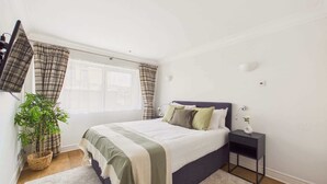 A bedroom with a bed, a nightstand, a lamp, a potted plant, and a window with curtains.