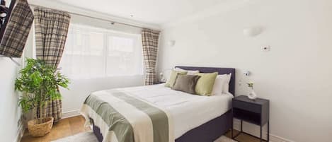 A bedroom with a bed, a nightstand, a lamp, a potted plant, and a window with curtains.