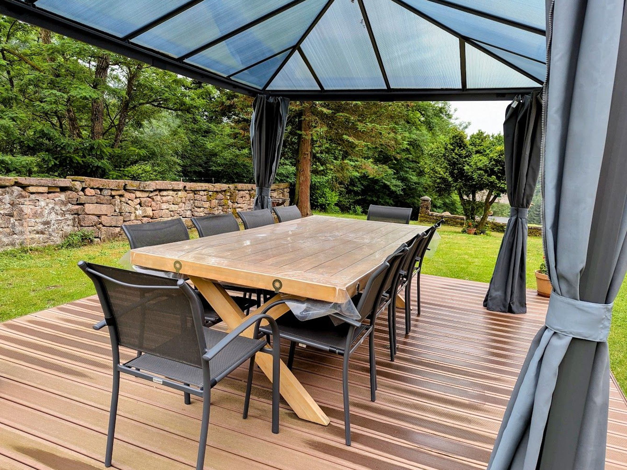 Un patio al aire libre con una mesa y sillas de madera, una cubierta de vidrio y un muro de piedra de fondo.
