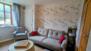 Una sala de estar con un sofá gris, un sillón más pequeño, una mesa de centro redonda con un tablero de ajedrez y una pared con diseño de ladrillo.
