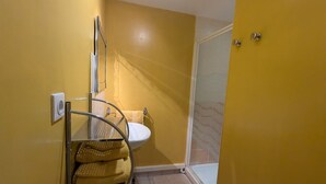 Un baño con una pared amarilla, un lavamanos blanco, una cabina de ducha de vidrio y un perchero con toallas.