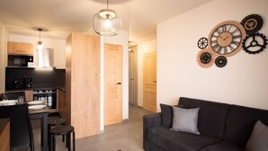 A modern kitchen with wooden cabinets, a dining area with a table and chairs, a grey sofa, and a wall clock with gears. - New T2 Apartment with balcony, covered parking, 4 guests. (Valmorel)