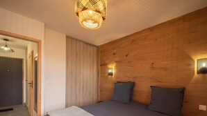 A bedroom with a wooden headboard, a bed with grey bedding, a bedside table, and a wall-mounted light fixture. - New T2 Apartment with balcony, covered parking, 4 guests. (Valmorel)