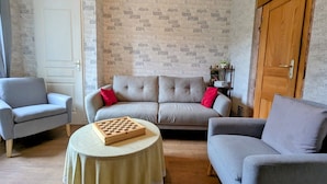 A living room with a grey sofa, two armchairs, a small round table with a chessboard, a brick-patterned wall, and a wooden door. - Iris Room in renovated house amidst nature (Moyenmoutier)