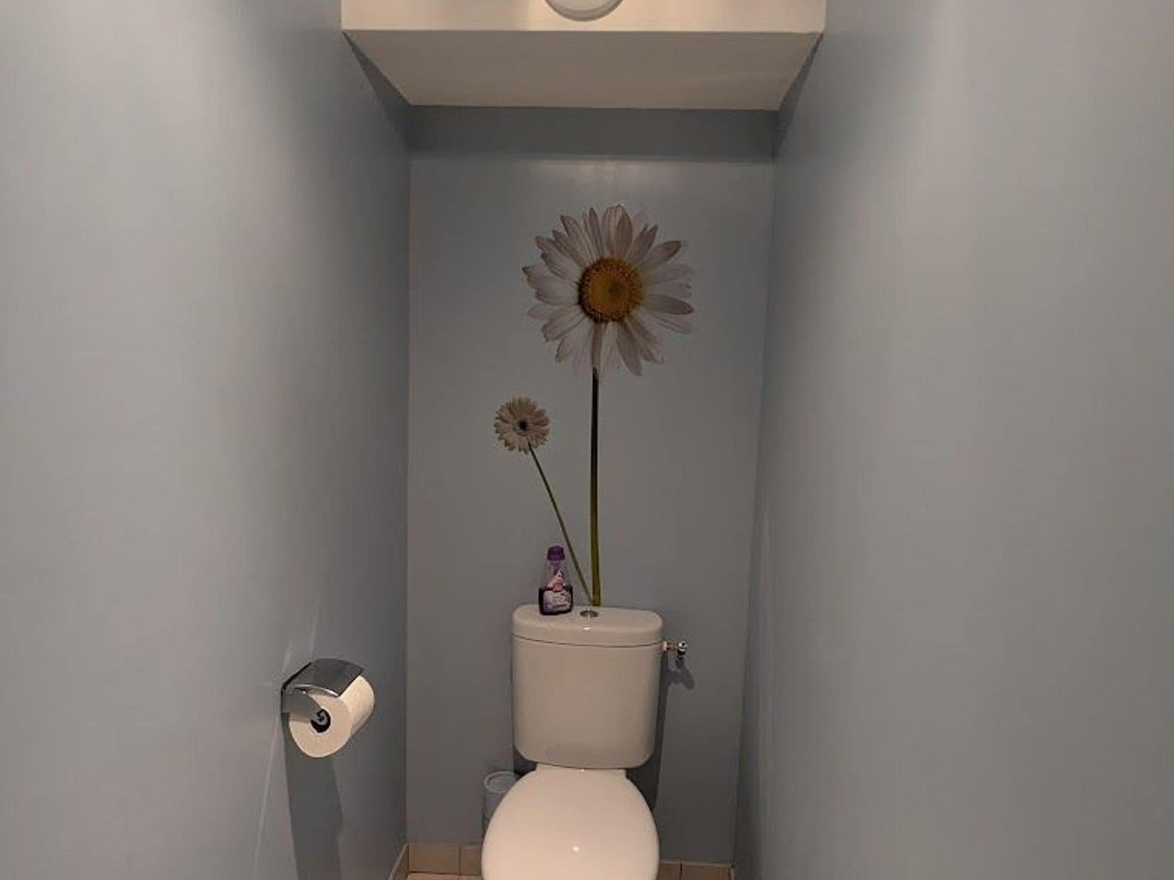 Une salle de bain avec des toilettes, un rouleau de papier toilette et une seule fleur posée sur le réservoir des toilettes.