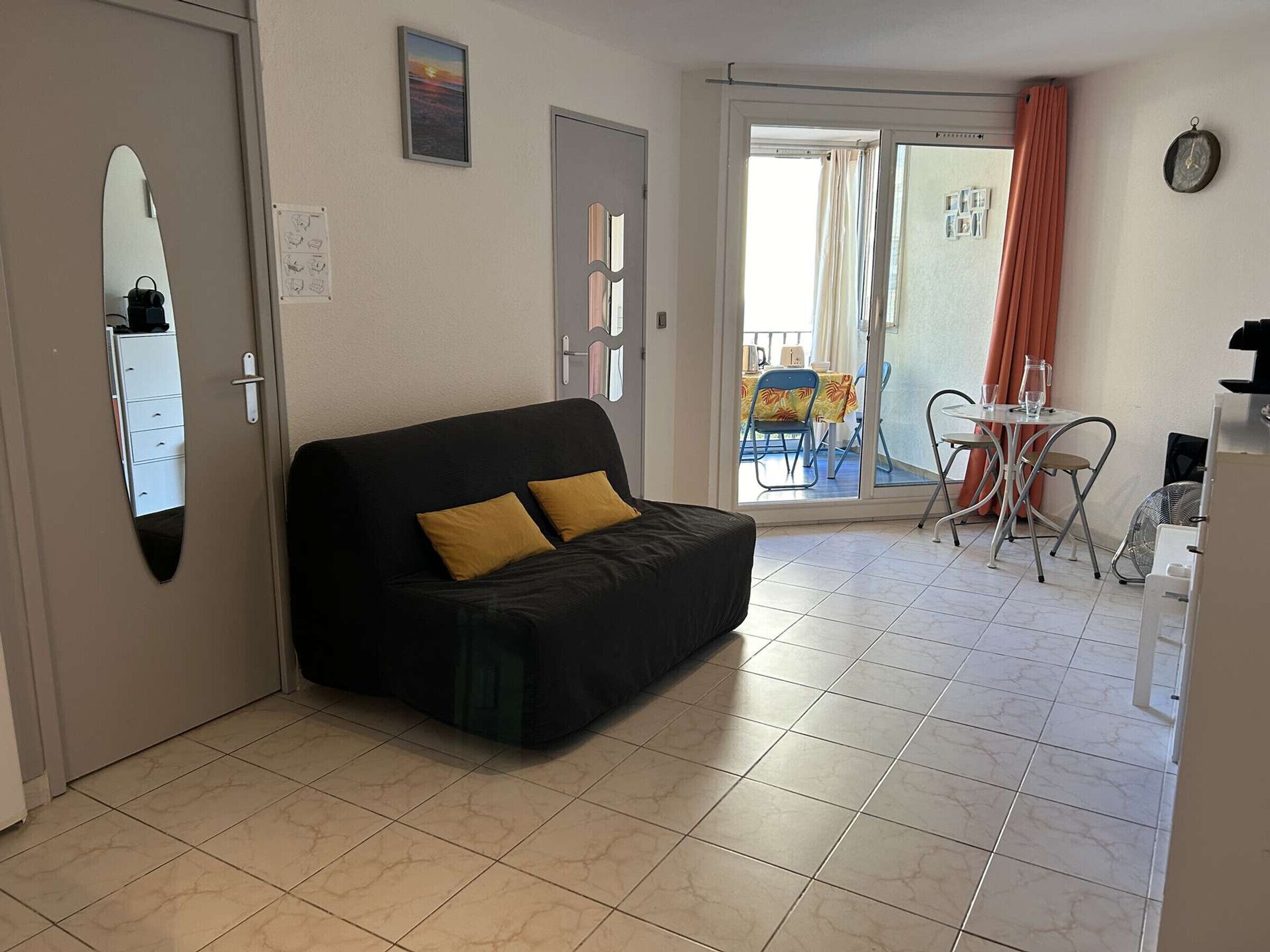Un salon avec un canapé noir, une petite table entourée de chaises, un miroir et une porte-fenêtre coulissante donnant sur un balcon.